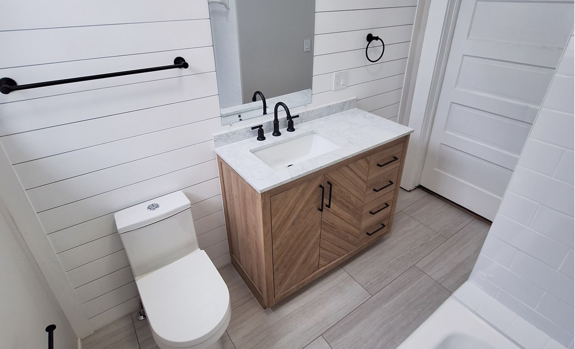 Bright bathroom with white shiplap walls, wood vanity, toilet, and black towel bar.