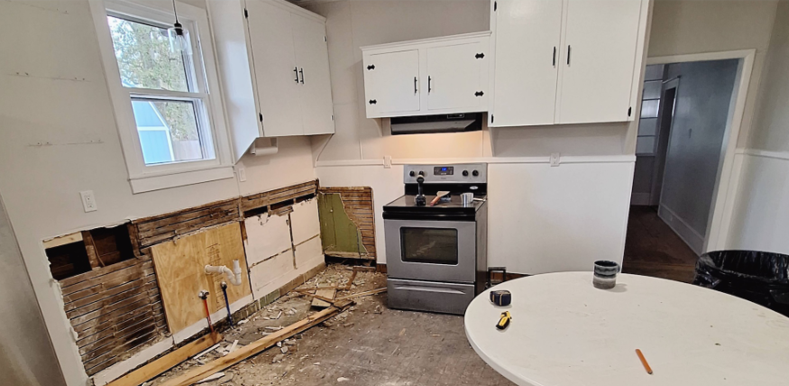 Damaged kitchen with exposed wall studs, missing cabinets, and a stove beneath white upper cabinets