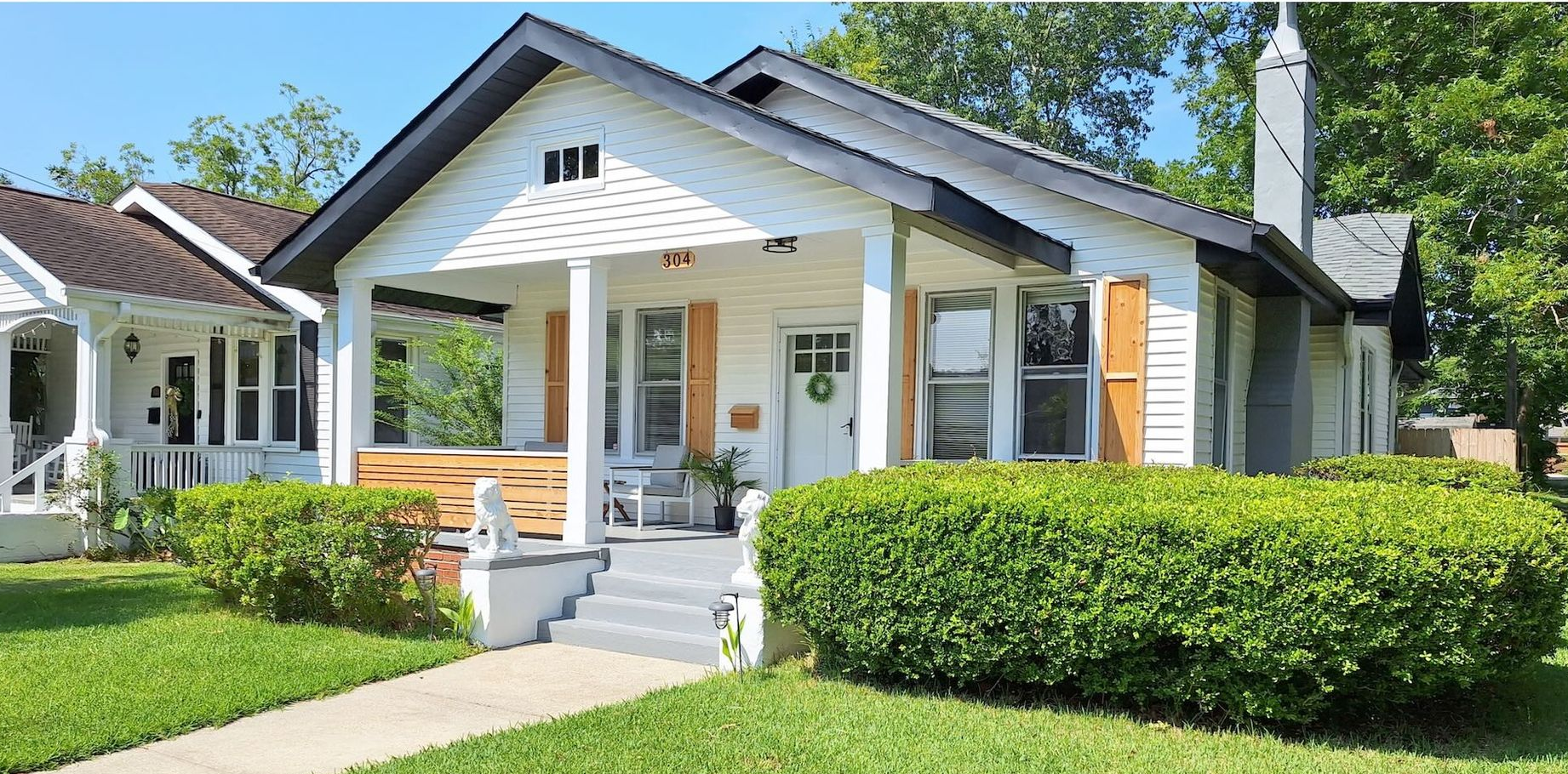 Single-story white house with a front porch, green hedges, and a manicured lawn in bright sunlight