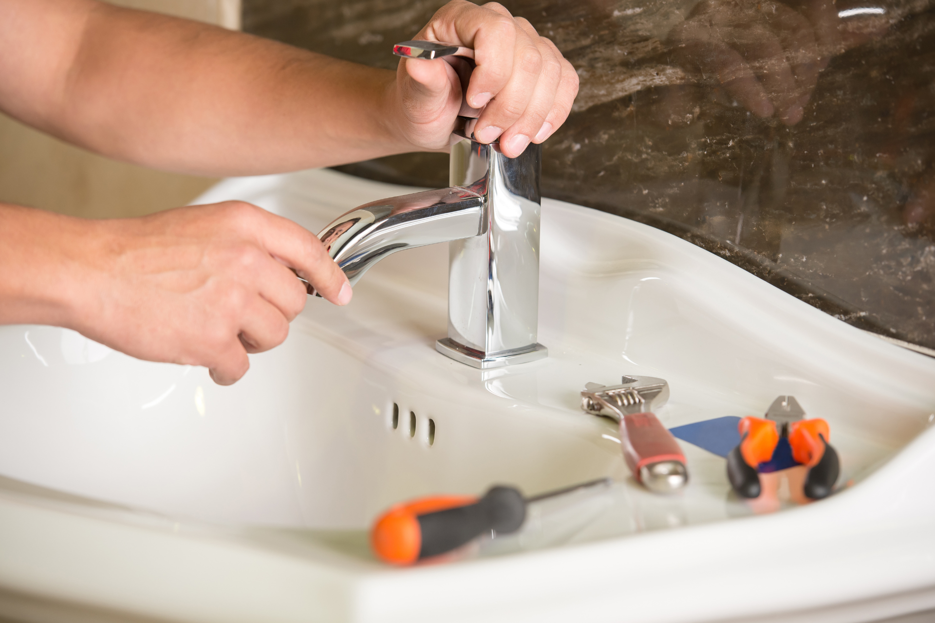 Hands fixing a chrome bathroom sink faucet with tools on the basin.