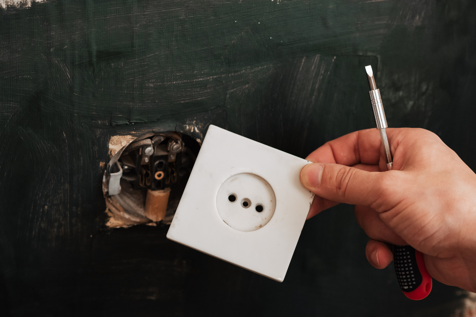 Hand holding screwdriver and outlet cover beside exposed electrical box on a wall