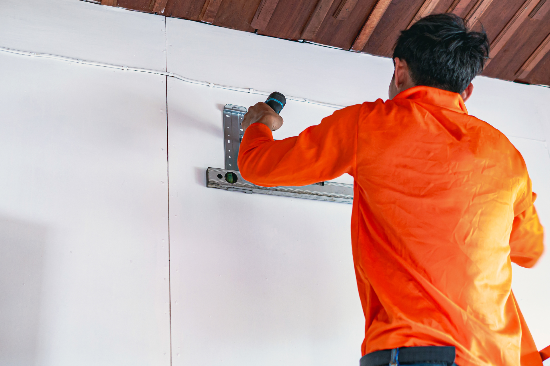 Worker in an orange shirt installing a wall-mounted device indoors.