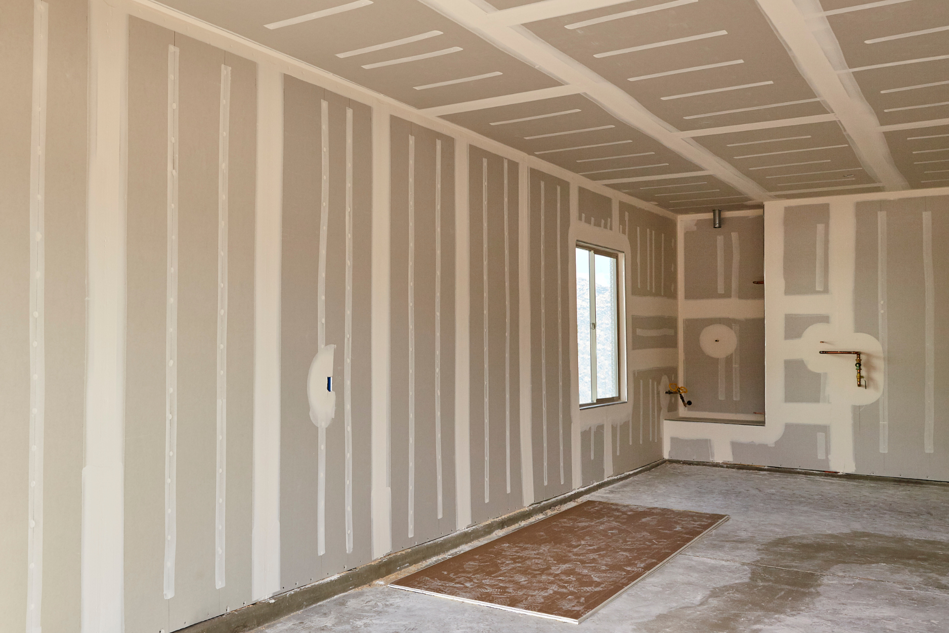 Unfinished drywall room with taped seams, window, and a carpet patch on the floor