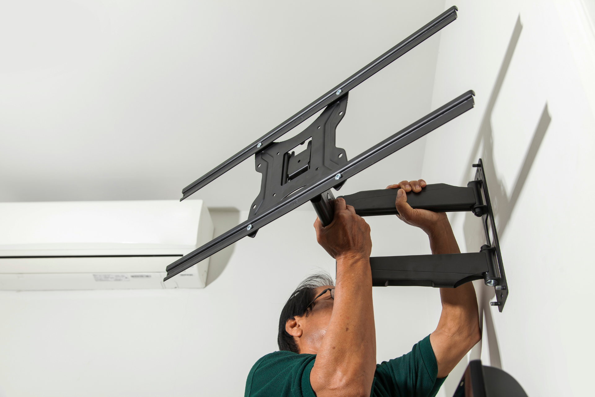 Person mounting a wall bracket for a TV, holding a large flat-screen display above their head