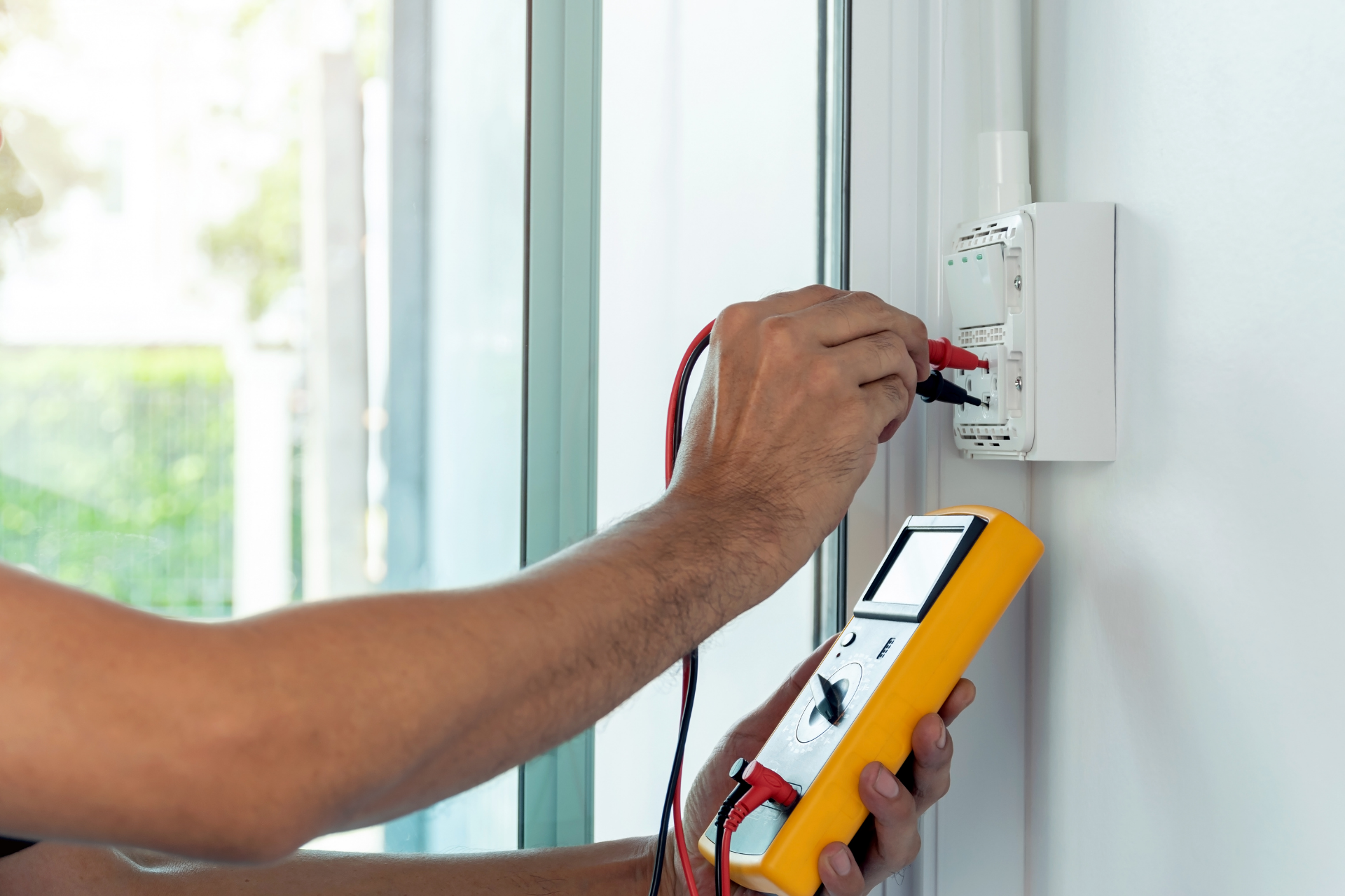 Technician testing wall outlet wiring with a multimeter inside a bright room