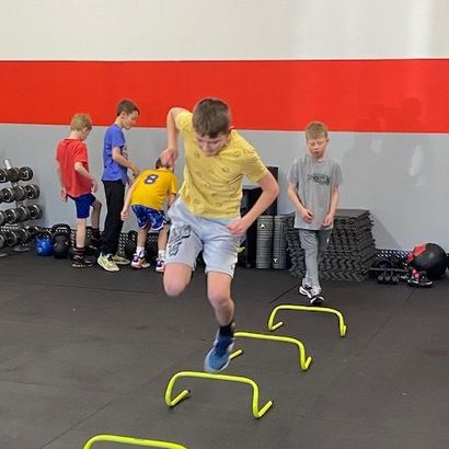 Boy jumping hurdles in a gym, others watching. Yellow hurdles, red wall, exercise equipment in background.
