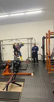 Person lifting weights in a gym with an instructor, large anime character on wall.