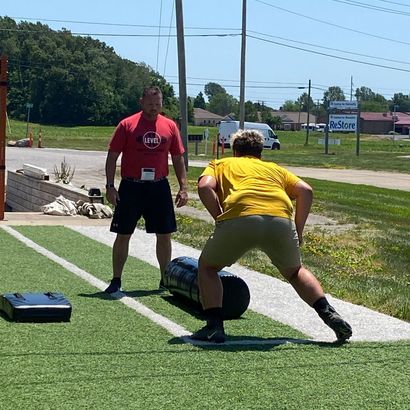 Man observing another man doing a sled drag exercise on artificial turf.