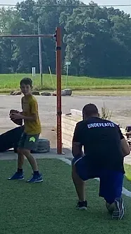Boy holding a football, practicing with coach outdoors. Green turf, orange pole, sunny day.