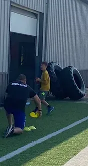 Person in black shirt kneeling, coaching boy holding ball; tires in background. Outdoor training area.