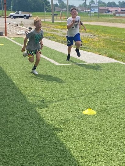 Two boys running on artificial turf. One wears a green shirt, the other a white shirt, both running towards the viewer.