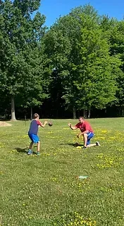 Boy throwing ball to man kneeling on grassy field with trees.