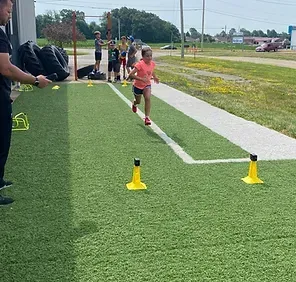 Girl running on artificial turf, guided by a coach. Other children and equipment are in the background.