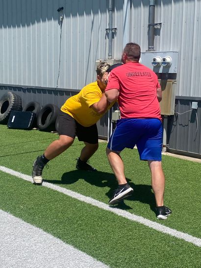 Man in red shirt coaches person in yellow shirt on a field; they are in a tackling stance.