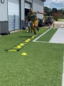 Person jumping through agility ladder on green turf outdoors.
