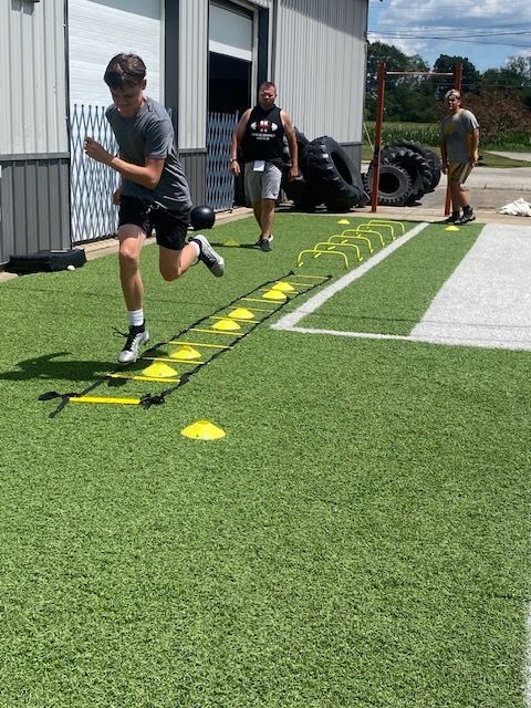 Person running agility ladder drill on artificial turf with observers.