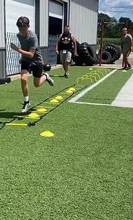 Teenager running through an agility ladder, followed by others on a turf field near tires and a building.