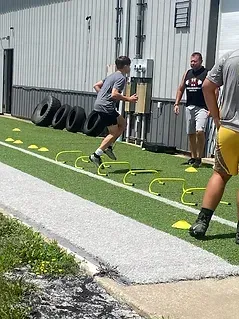 A young person running agility drills over small hurdles on a green turf field, observed by two others.