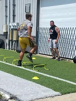 Athlete jumping over hurdles on a green turf field, being coached by a man.