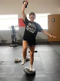 Woman balancing on a dome, holding kettlebell overhead. Gym setting, other leg raised, arms outstretched.