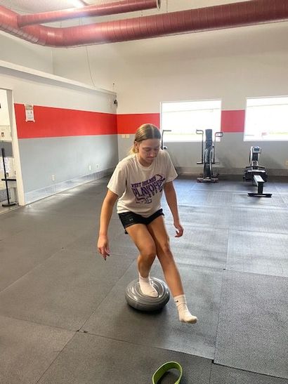 Woman balances on a BOSU ball in a gym setting, lifting one leg, wearing athletic clothing.