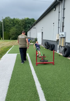 A person pulls a weighted sled on artificial turf, with another person walking behind.