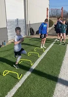 Boy running through small hurdles on green turf, other people watching. Outdoors.