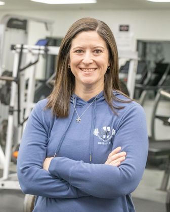 Woman with arms crossed, smiling in a gym, wearing a blue hoodie.