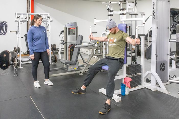 Man using a chest fly machine in a gym, with a woman observing. Black workout floor and gym equipment.