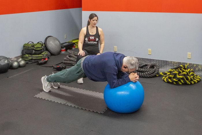 Man doing plank on exercise ball, supervised by woman in gym.