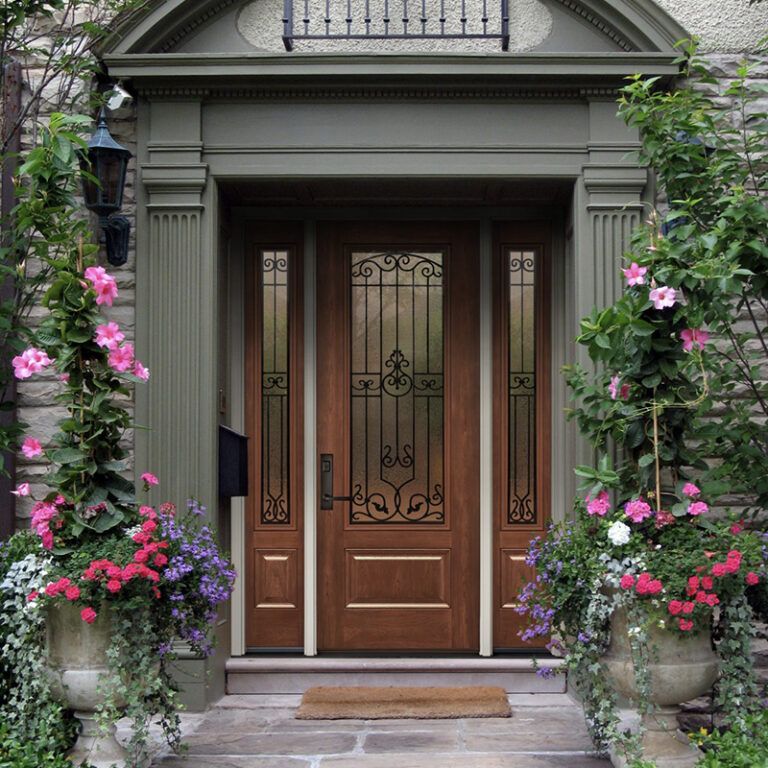 A wooden front door with sidelights and an ornate iron design. Flowers in pots flank the entrance.