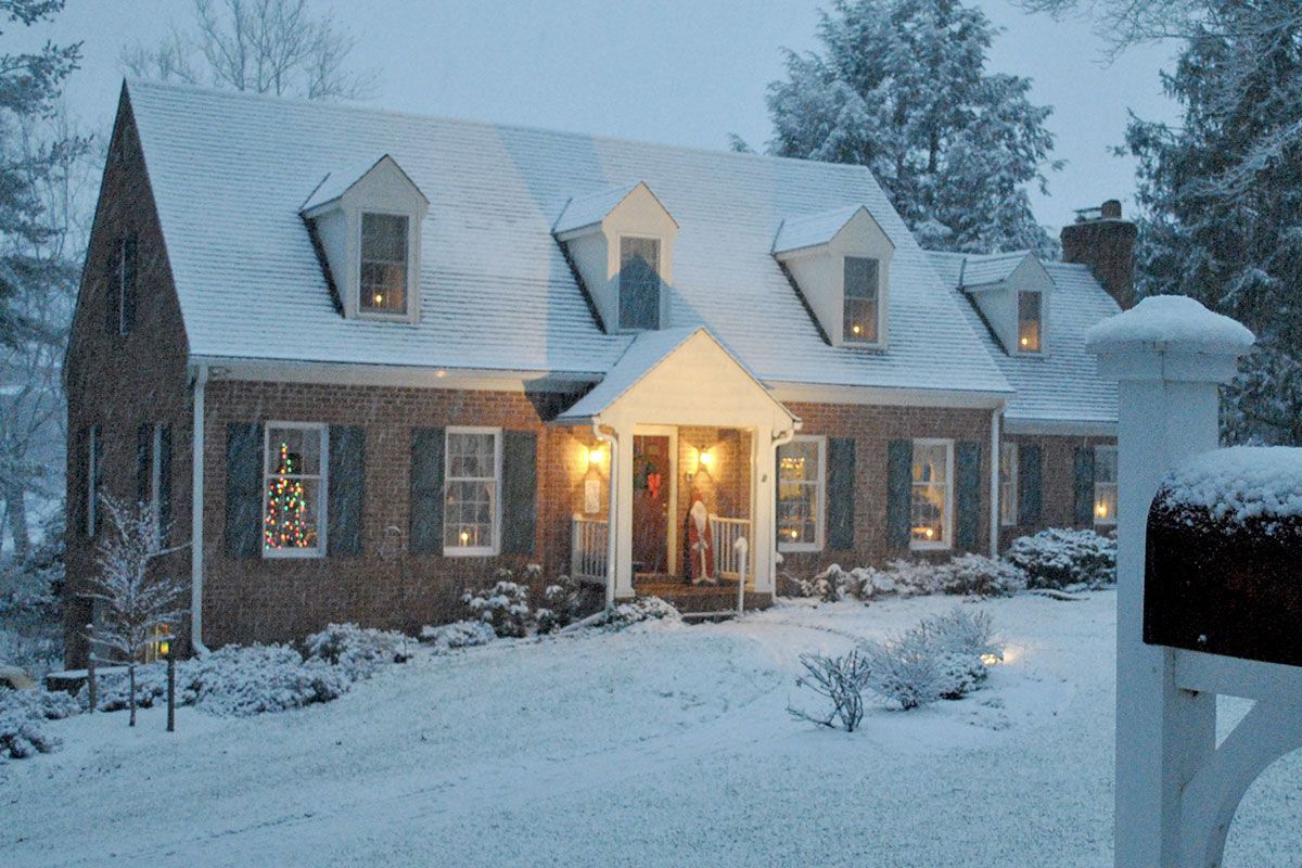 A snow-covered brick house at dusk with lights on inside. A mailbox and landscaping are also covered in snow.