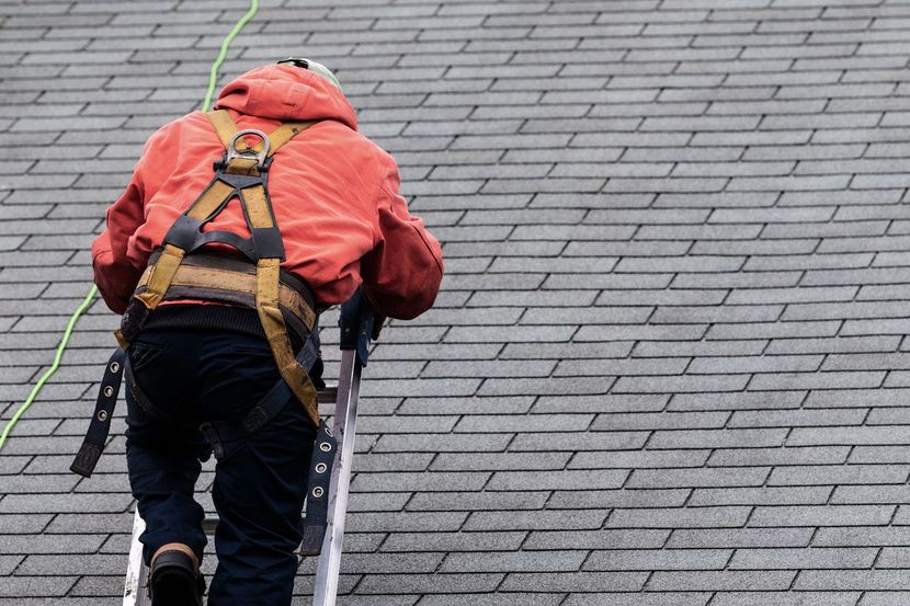 Century Home Improvements - Roofer in orange jacket, ascending a ladder on a gray shingle roof, wearing a safety harness.