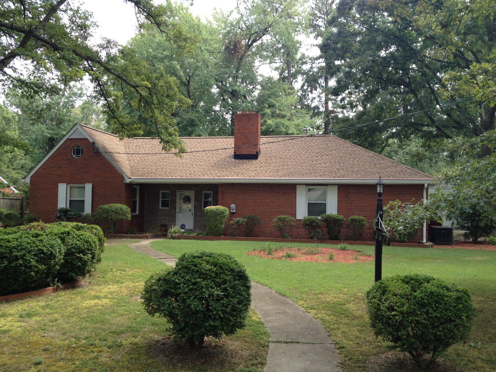 Red brick house with a brown roof, chimney, and a small front yard with shrubs and a walkway.