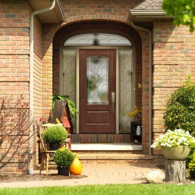 Brown door with glass panels within a brick arched entryway, decorated with plants, a wooden chair, and a pumpkin.