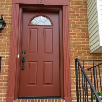 Red front door with arched window, brick exterior, and red trim. Black railing to the right.