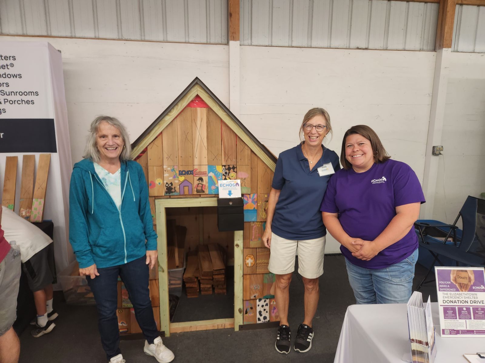Three women pose next to a miniature house, possibly at a fair or exhibit.