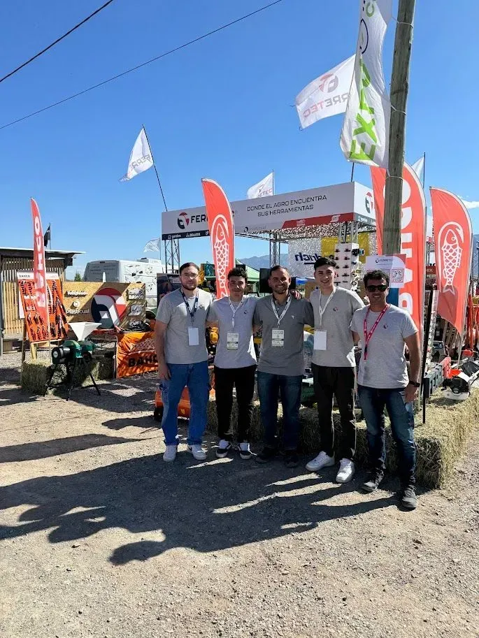 Cinco personas posando al aire libre en un stand de un evento con banderas rojas y carpas bajo un cielo azul despejado.