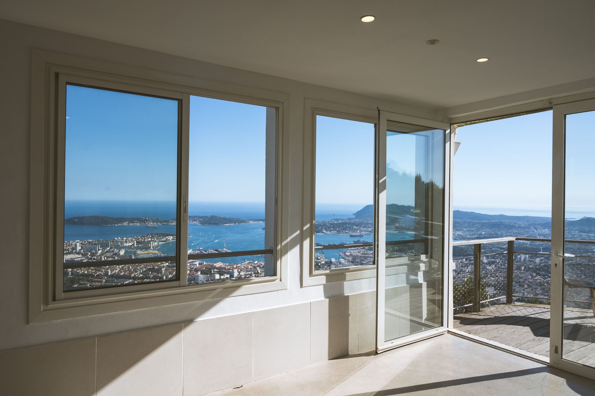Habitación luminosa con grandes ventanales y una puerta de cristal abierta con vistas a una ciudad costera y al océano.