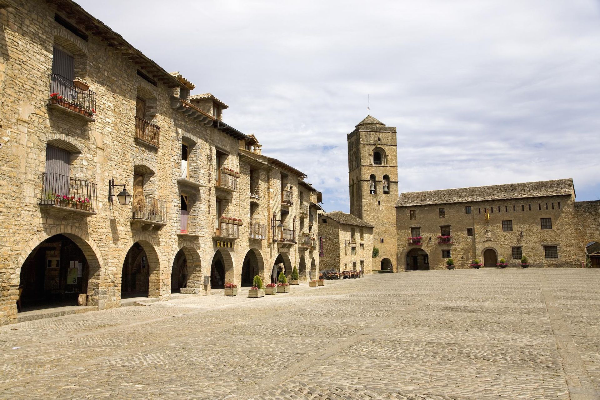 Plaza adoquinada bordeada de edificios de piedra; torre de la iglesia al fondo bajo un cielo nublado.