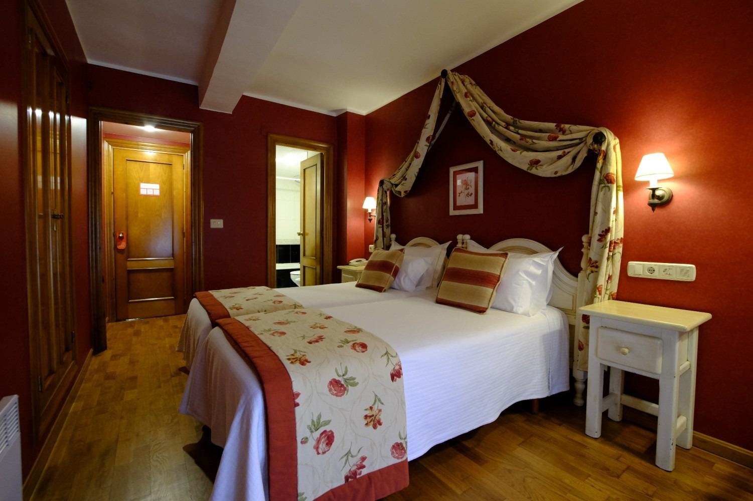 Habitación de hotel con dos camas individuales, paredes rojas, suelos de madera y baño visible.
