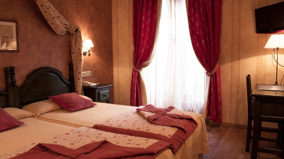 Habitación de hotel con dos camas, cortinas rojas, escritorio y lámpara.