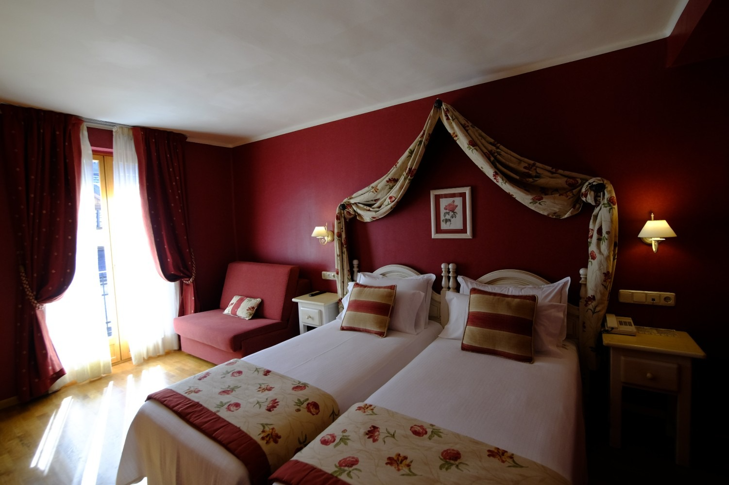 Habitación de hotel con dos camas individuales, paredes rojas, dosel y ventana con cortinas.