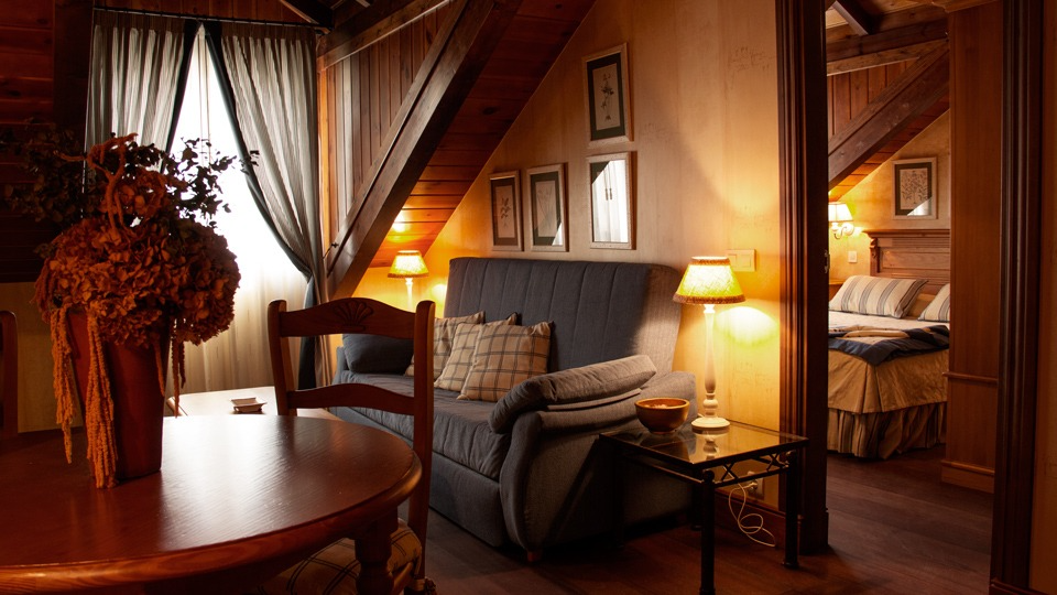 Acogedora habitación abuhardillada con sofá, mesa y cama visibles en la puerta; iluminación cálida.
