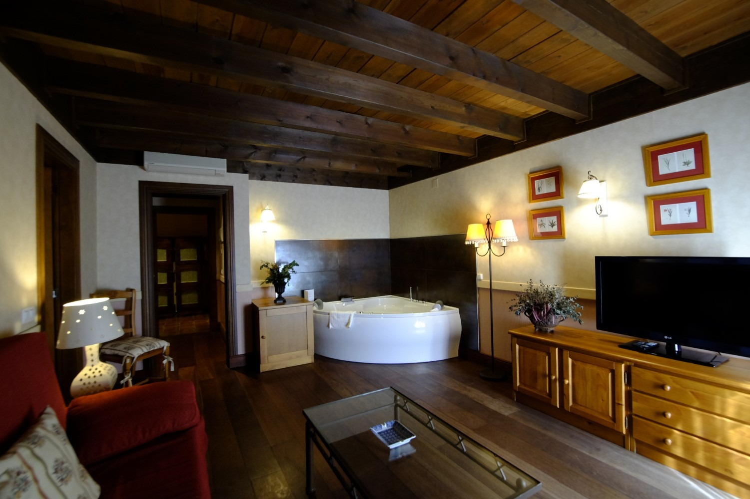 Acogedora habitación de hotel con vigas de madera, jacuzzi, sofá rojo, TV y muebles de madera.