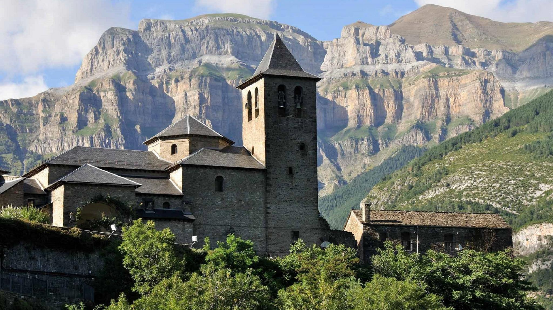 Iglesia de piedra con campanario, enclavada en un valle con montañas al fondo.