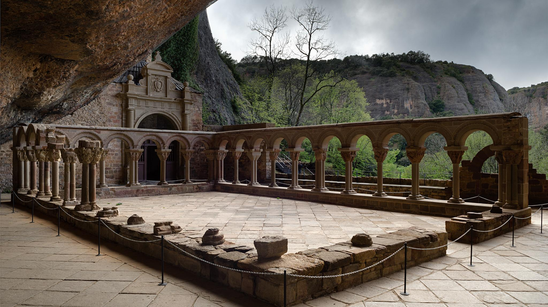 Claustro de piedra con pasarelas arqueadas, situado bajo un acantilado, con árboles al fondo.