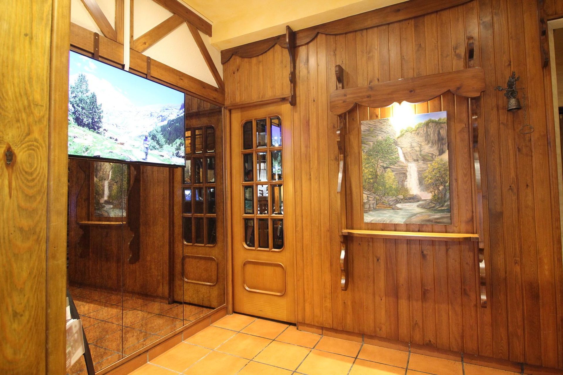 Interior revestido de madera con puerta, dos cuadros de paisajes enmarcados y un espejo.