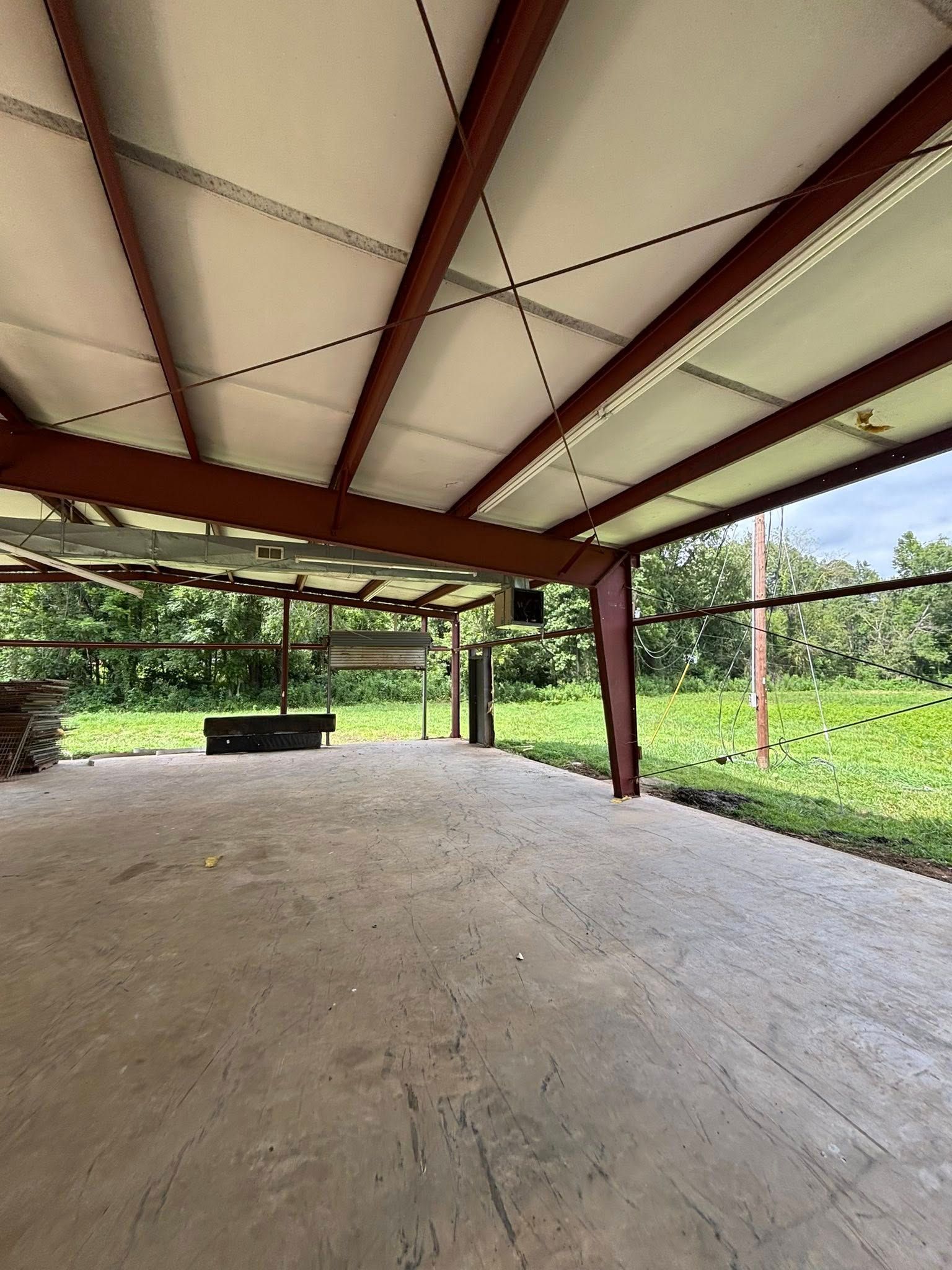 Open-air concrete pavilion with a metal roof, red beams, and grassy yard beyond