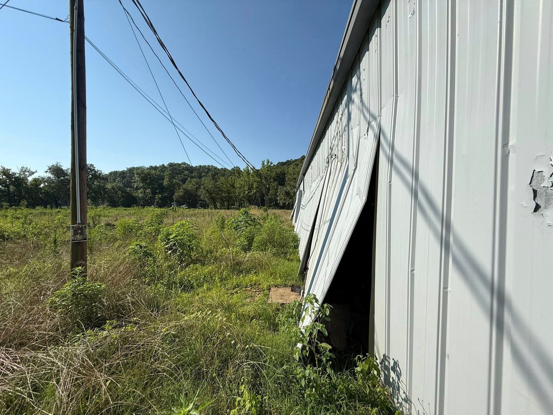 White metal building with a bent side panel beside a grassy field and power lines under a blue sky